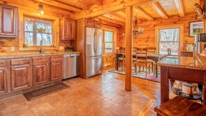 Une cuisine rustique aménagée dans une cabane en rondins, avec des meubles de rangement en bois, un réfrigérateur en acier inoxydable et un coin repas comprenant une table et des chaises en bois.