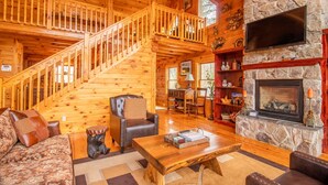 L'intérieur d'une cabane en rondins, avec un escalier en bois, une cheminée en pierre et un espace de vie chaleureux comprenant un canapé et une table basse.