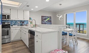 Uma cozinha moderna com armários brancos, forno embutido e lava-louças. Há uma ilha grande com uma pia, e uma área de jantar com mesa e cadeiras. Uma janela grande oferece vista para o oceano.