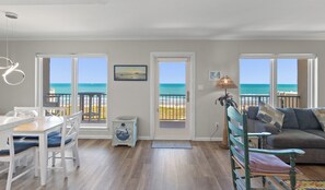 A room with a wooden floor, a dining table with chairs, a sofa, a lamp, and a view of the beach through a door.