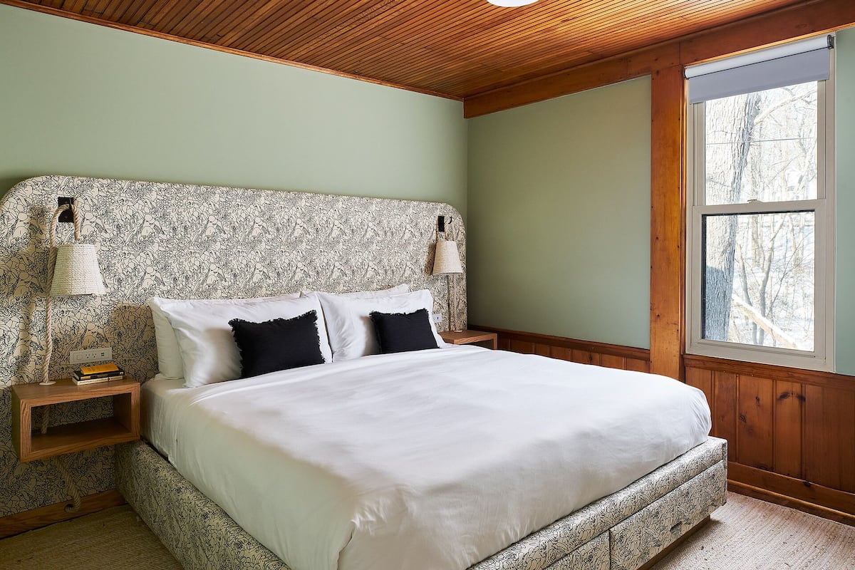 A bedroom with a wooden ceiling, a bed with white bedding, patterned headboard, bedside lamps, and a window with a view of trees.