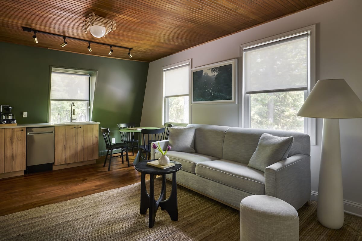 A modern living room with a grey sofa, a small round table with a vase, a tall floor lamp, and a kitchen area with wooden cabinets and a dishwasher.