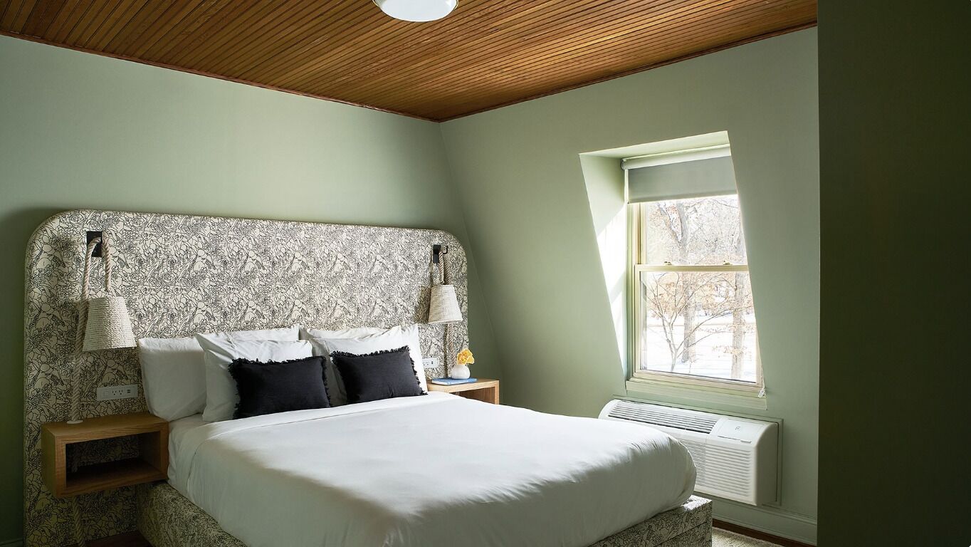 A bedroom with a patterned headboard, a bed with white linens, a nightstand with a lamp, and a window with a view of trees.