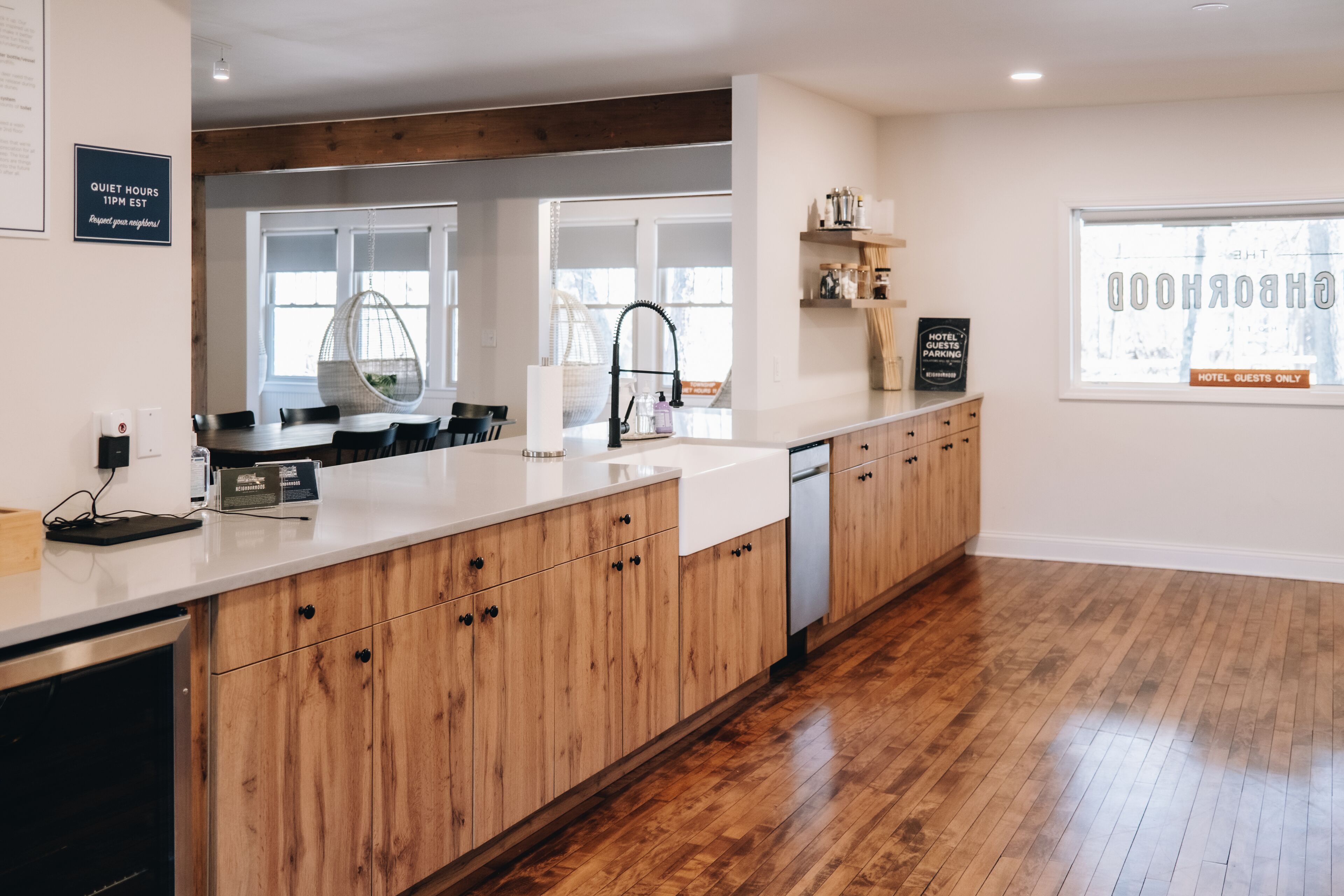 A modern kitchen with wooden cabinets, a white countertop, and a built-in dishwasher. There is a hanging chair near the window and a sign indicating quiet hours.