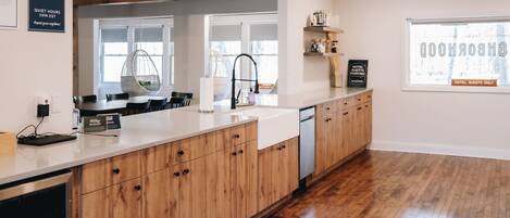 A modern kitchen with wooden cabinets, a white countertop, and a built-in dishwasher. There is a hanging chair near the window and a sign indicating quiet hours.