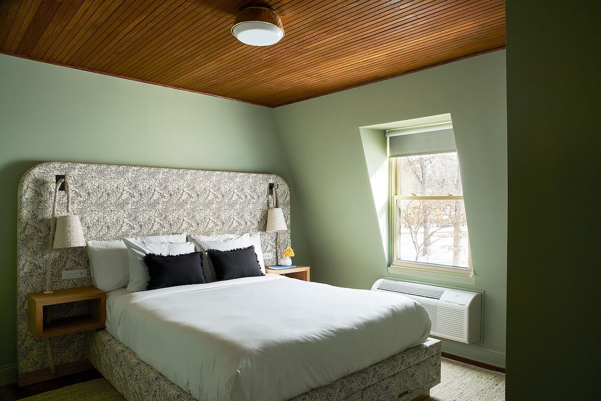 A bedroom with a wooden ceiling, a bed with white bedding, a nightstand with a lamp, and a window with a view of trees.