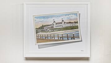 A framed illustration of a hotel with a waterfront view, including a pier and a large building with multiple flags.