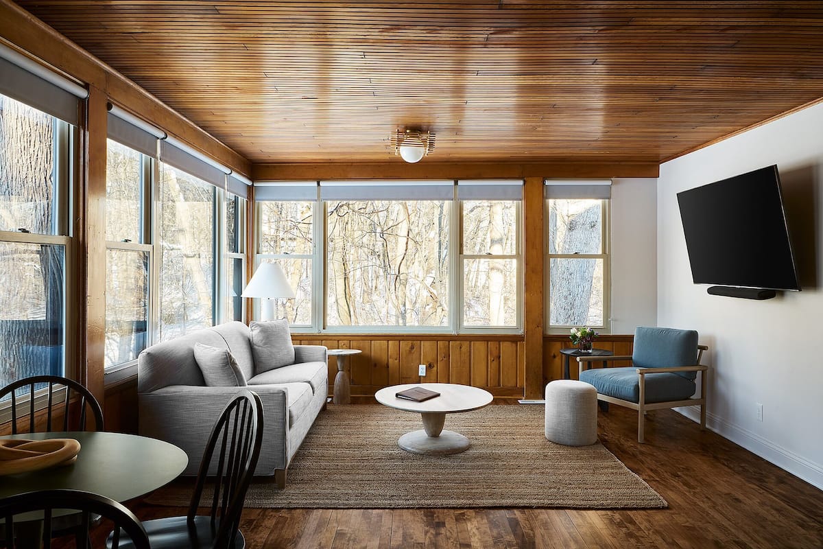 A spacious living room with a wooden ceiling, large windows with a view of trees, a flat-screen TV mounted on the wall, and a round coffee table.