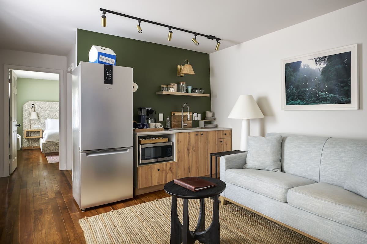 A compact kitchen with a refrigerator, microwave, and coffee maker. There is a wooden cabinet and a small table with a book. A gray sofa is in the living area with a painting on the wall.