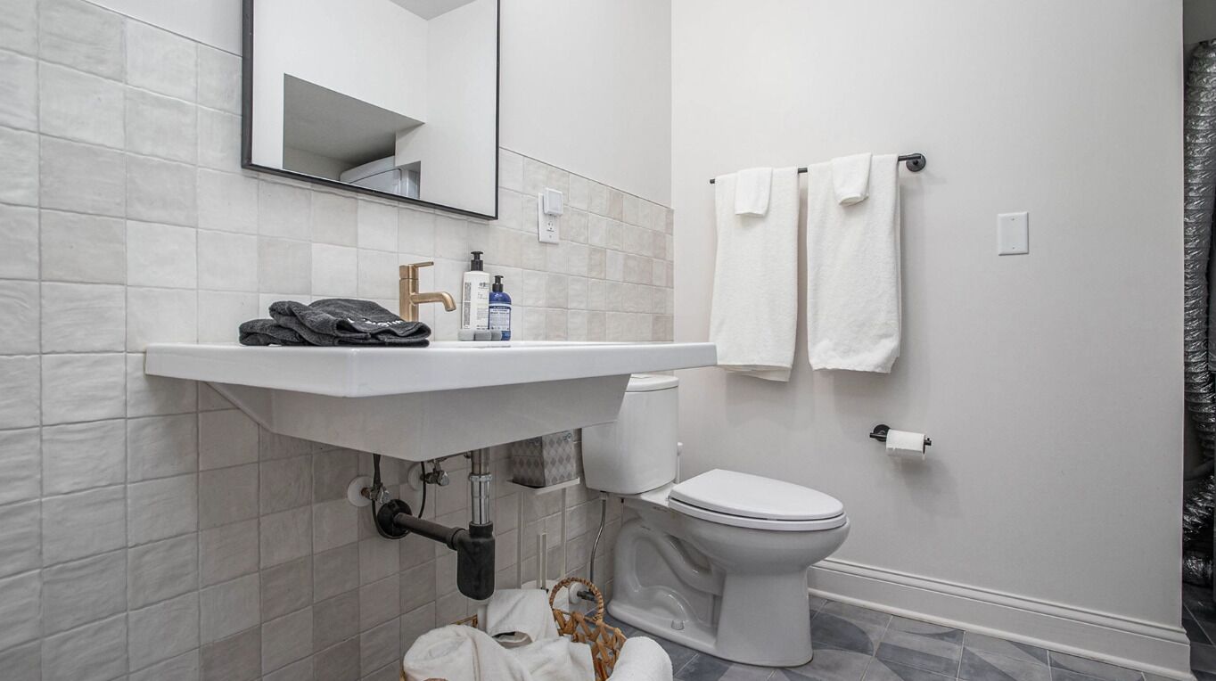 A modern bathroom with a white sink, a toilet, and a mirror. There are towels hanging on a rack and a basket with toiletries on the floor.