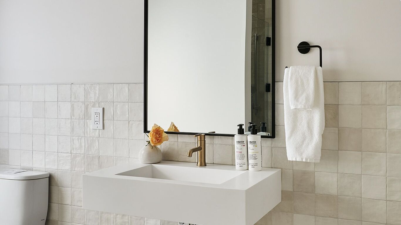 A modern bathroom with a white sink, a gold faucet, and a mirror above it. There are two white towels hanging on a towel rack, and a vase with a single flower on the sink counter.