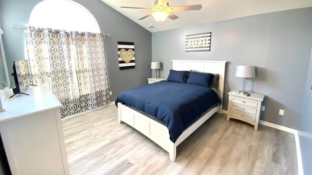 Un dormitorio con cama, mesitas de noche, ventilador de techo y ventana con cortina.