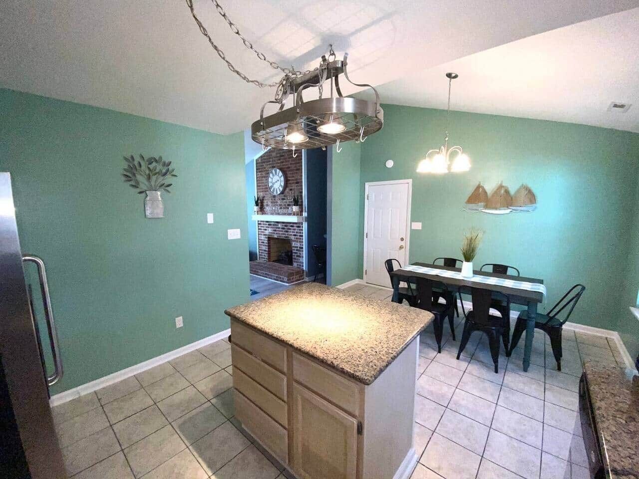 A kitchen with a central island, a dining table with chairs, a hanging light fixture, and a door leading to another room.