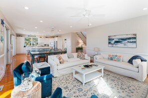 Amplia sala de estar con sofá blanco, sillón azul y una mesa de centro. Una escalera conduce al piso superior, y se observa una cocina al fondo.