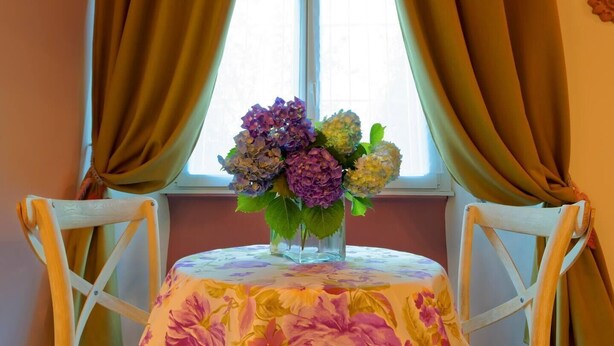 Une table recouverte d’une nappe à motifs floraux, un vase contenant des fleurs violettes et jaunes, et une fenêtre avec des rideaux.