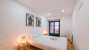 A bedroom with a bed, two bedside lamps, a wooden nightstand, a window with a view, and two framed pictures on the wall. - GuestReady - Green Bay Garden (Porto)