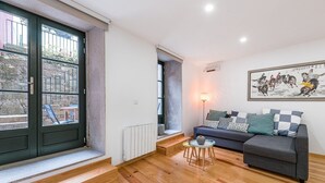 A modern living room with a grey sofa, patterned cushions, a small round table, and a large window with a view of a stone staircase. - GuestReady - Green Bay Garden (Porto)