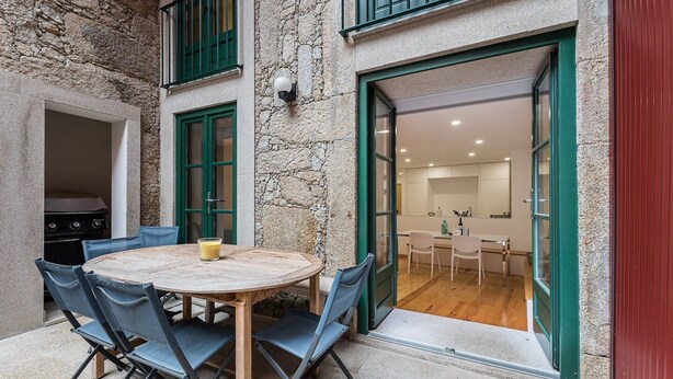 A stone building with a round wooden table and blue chairs, a grill, and a kitchen visible through an open door. - GuestReady - Green Bay Garden (Porto)