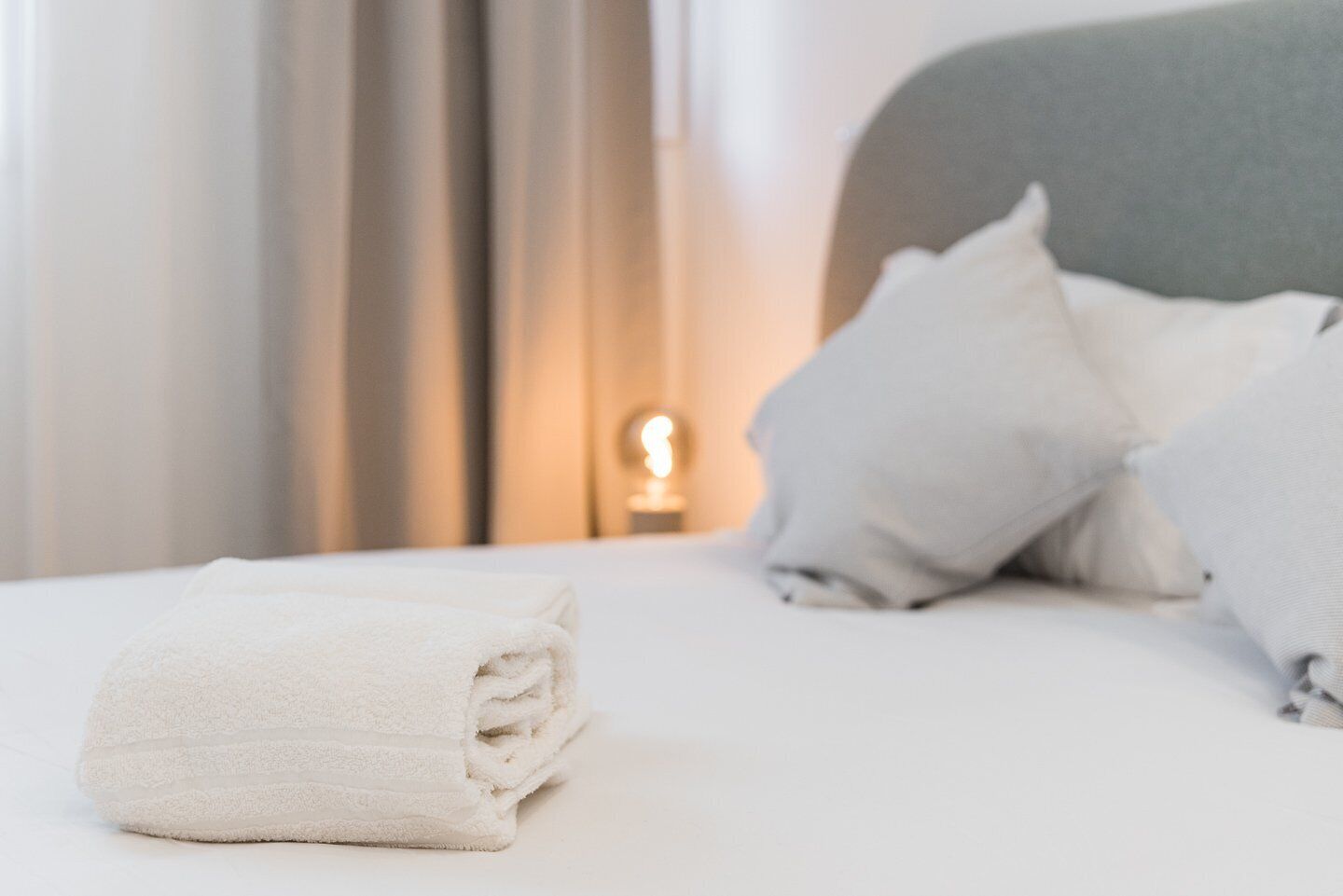 A neatly made bed with white linens and pillows, a bedside lamp, and a curtain in a hotel room.