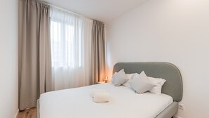 Ein ordentlich bezogenes Bett mit grauem Kopfteil, weißer Bettwäsche und einer Nachttischlampe.