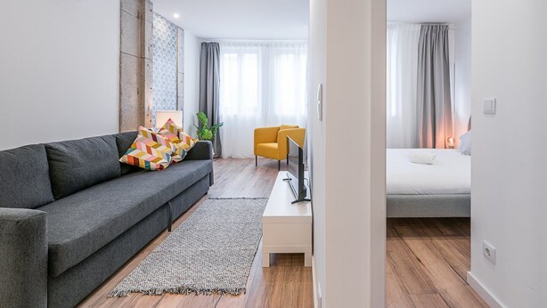 Ein modernes Wohnzimmer mit einem grauen Sofa, einem gelben Sessel und einem Fernseher auf einem weißen Couchtisch. Im Hintergrund ist ein Bett mit weißer Bettwäsche zu sehen.