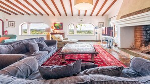 Amplia sala de estar con un gran sofá, una mesa de centro y una chimenea. Varias ventanas ofrecen vistas al exterior.