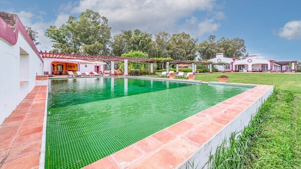 Una piscina con borde de azulejos, rodeada por un edificio blanco con techo rojo y una pérgola con sillas de descanso.