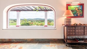 Una habitación con un ventanal arqueado que da a un paisaje, un aparador de madera con cajones y un cuadro enmarcado en la pared.