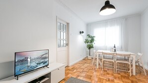 A modern living room with a flat-screen TV, wooden flooring, a dining table with chairs, a potted plant, and a pendant light.