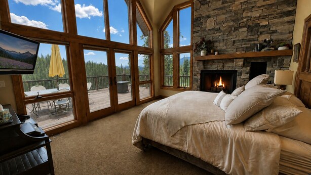 A bedroom with a large bed, a stone fireplace, and a balcony with a view of trees and mountains. - Purgatory Cabin with Views and Hot Tub (Durango)