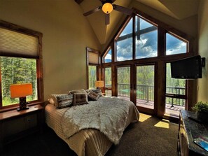 Relax in this serene bedroom with a large bed, ceiling fan, and flat-screen TV, plus a balcony with a view of the surrounding greenery.