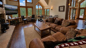 A spacious living room with a large couch, a coffee table, and a TV mounted on a stone wall. There are multiple windows providing natural light and views of the outdoors. - Purgatory Cabin with Views and Hot Tub (Durango)
