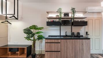 A compact kitchen with a black countertop, wooden cabinets, and a microwave. There are two potted plants, a hanging light, and a small shelf with kitchen items.