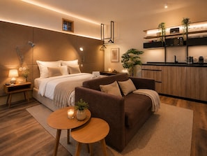A modern hotel room with a bed, sofa, bedside tables, a small kitchen area, and a dining table.