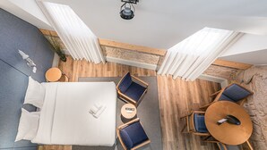 A modern hotel room with a large bed, a round dining table, and two chairs. There is a small round table with a book and a remote control. The room has wooden flooring and a stone accent wall. - GuestReady - Mercadores Apartments - Room G (Porto)