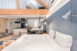 A modern hotel room with a large bed, a desk, a chair, a small table, a mirror, and a kitchenette.