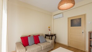 A living room with a beige sofa, red and checkered cushions, a small table with a vase, a white coffee table, and a wooden door with a textured panel. - GuestReady - City & Beach Garden Home (Porto)