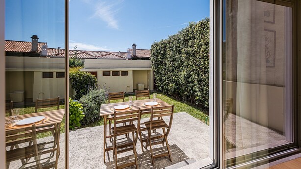 A patio with a table and chairs, a hedge, and a building with a tiled roof in the background. - GuestReady - City & Beach Garden Home (Porto)