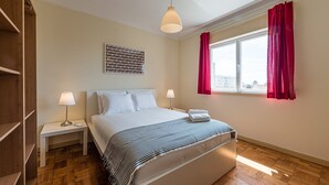 A hotel room with a bed, two bedside tables, a lamp on each table, a window with red curtains, and a city view. - GuestReady - City & Beach Garden Home (Porto)