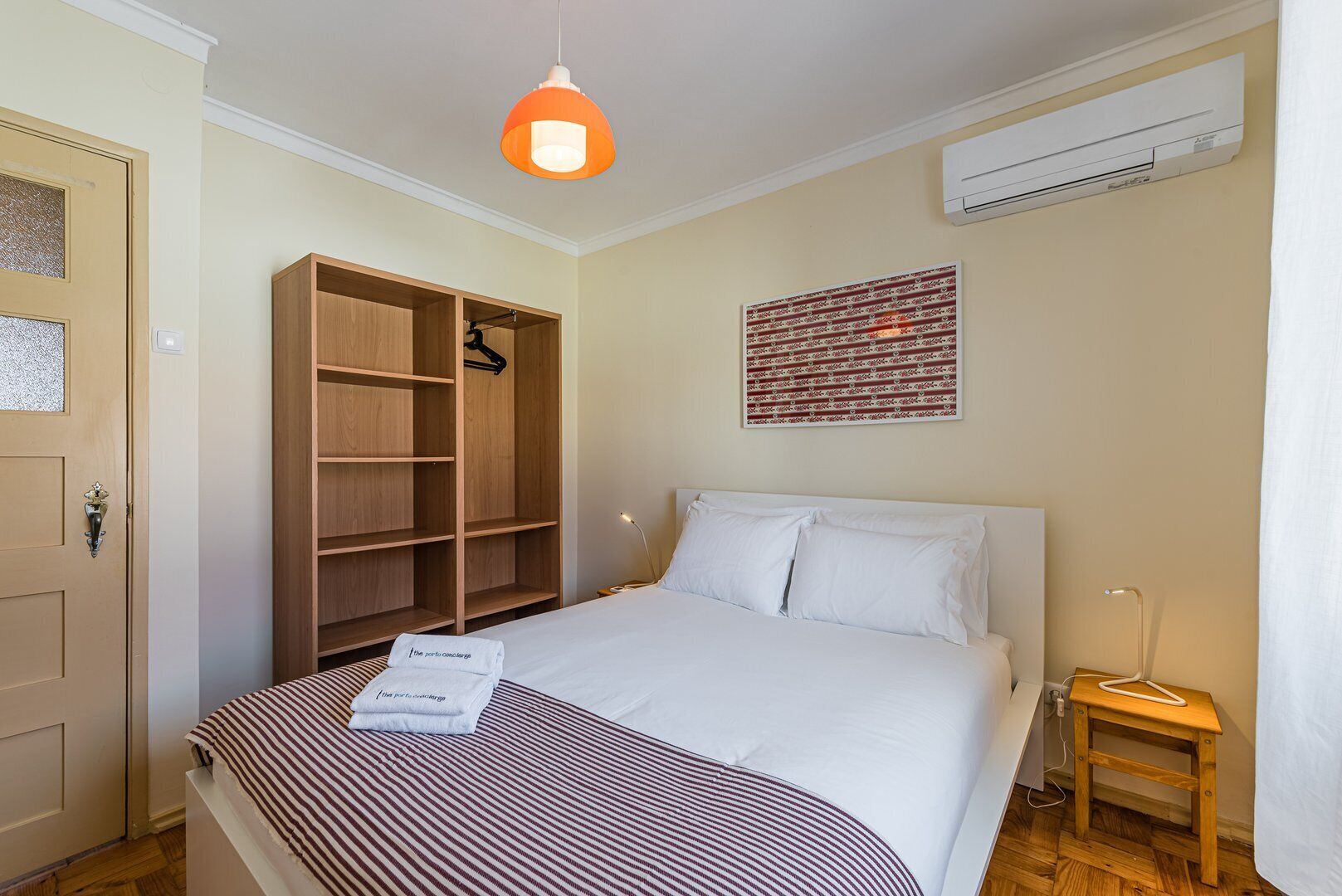 A bedroom with a bed, a wooden wardrobe, a hanging light fixture, an air conditioner, and a wall-mounted artwork.