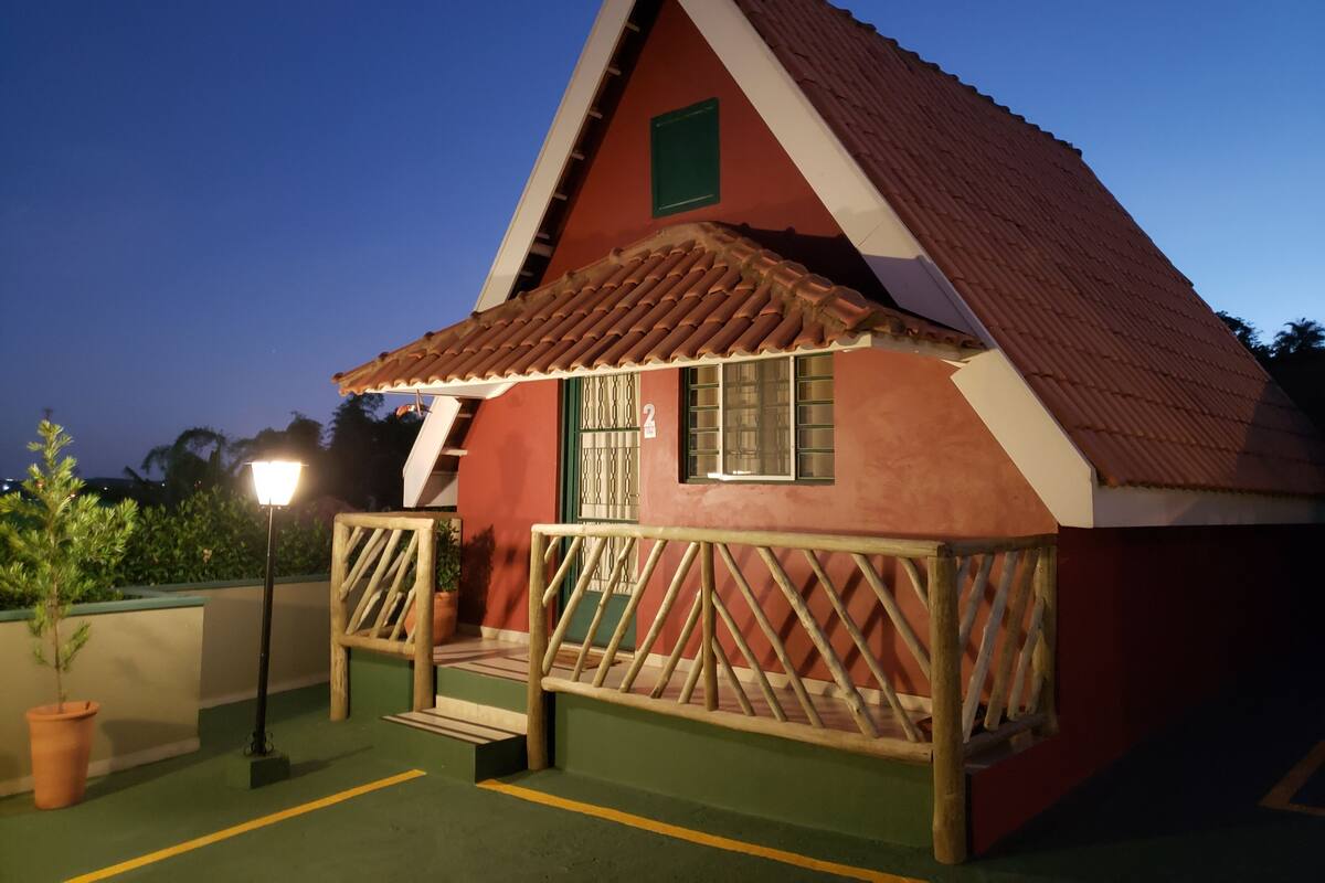 Une petite maison de plain-pied avec un toit rouge et des encadrements de fenêtres verts, dotée d’un porche en bois et d’un petit escalier menant à l’entrée.