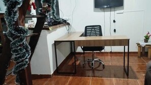 Una habitación con un escritorio de madera, una silla de oficina negra, una mesita con una planta y un televisor fijado a la pared.