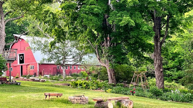 A red barn with a black door and a heart-shaped cutout, surrounded by lush greenery and trees. - Charming, updated 3 bedroom farmhouse farm stay.  (Williamsburg)