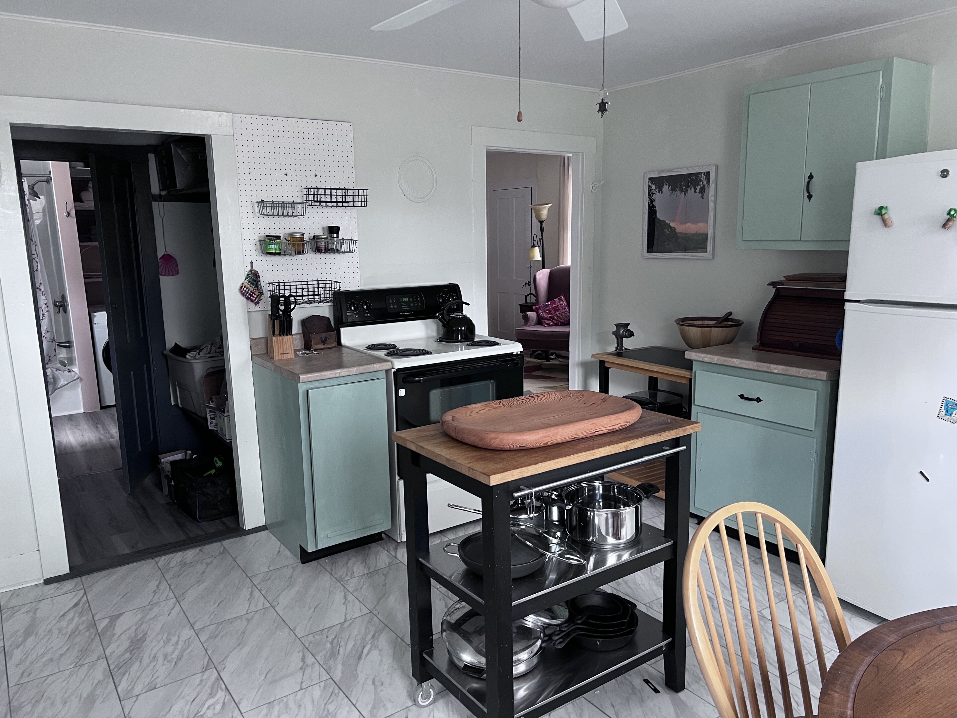 Une cuisine compacte équipée d’une cuisinière, d’un évier et de placards. On y trouve une table en bois avec une planche à découper et des casseroles. Une chaise est visible au premier plan.