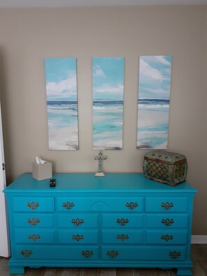 Een turquoise dressoir met versierde handgrepen, een afstandsbediening en een decoratieve doos. Daarboven hangen drie verticale schilderijen die een strandtafereel voorstellen.