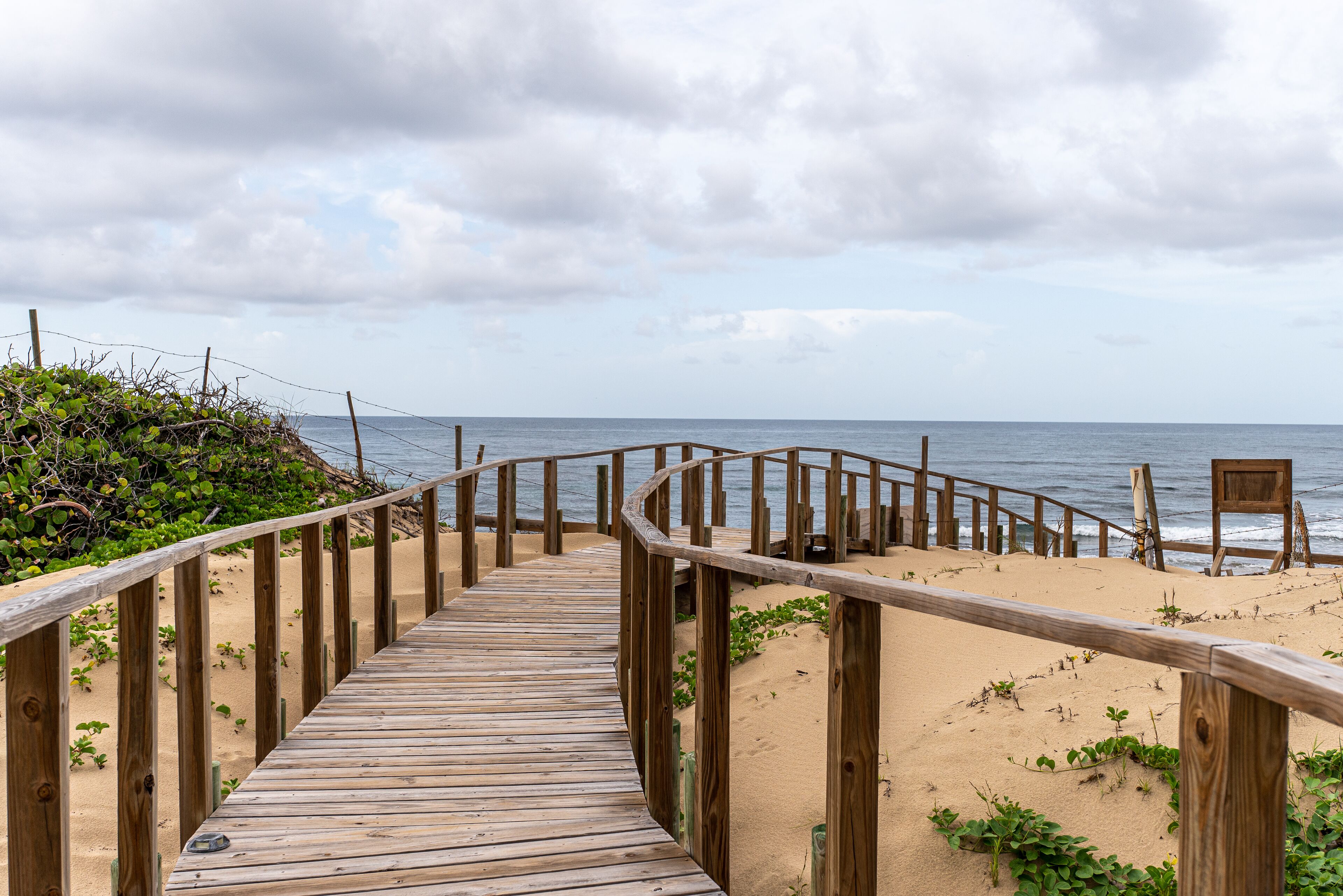 Ein hölzerner Steg führt zu einem Strand mit sandigem Ufer und bewölktem Himmel.