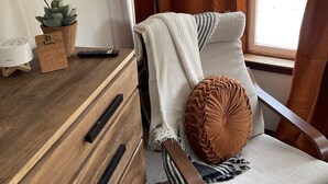 Un salon chaleureux avec une commode en bois, un fauteuil recouvert d’un plaid, une lampe sur pied et une plante en pot.