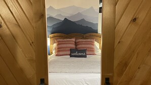 Un chambranle de porte en bois mène à un lit orné d’une fresque représentant une montagne, sur lequel est posé un coussin avec l’inscription « bienvenue », et un tapis à motifs.
