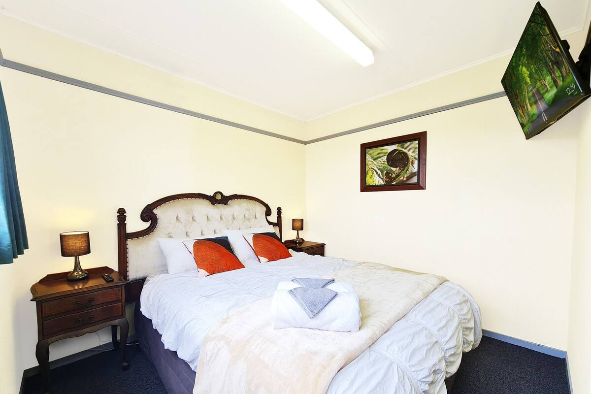 A neatly made bed with a white comforter and two orange pillows, a wooden headboard, and a small bedside table with a lamp.