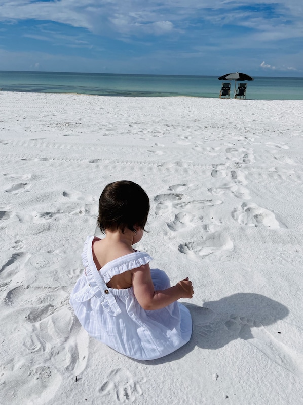 A child sitting on a sandy beach with footprints in the sand, a clear sky, and a distant umbrella.